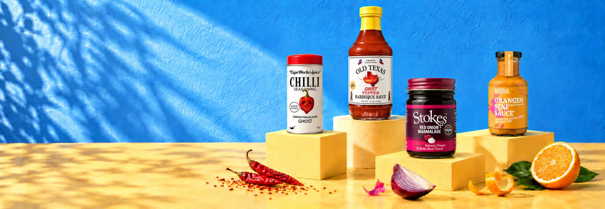 Cape Herb Ghost Chilli Rub, Old Texas Ghost Pepper BBQ-Sauce, Stokes Red Onion Marmelade und BSC Orangen-Senf-Sauce auf gelben Podesten vor einer blauen Wand, davor getrocknete Chilis, eine rote Zwiebel und eine Orange