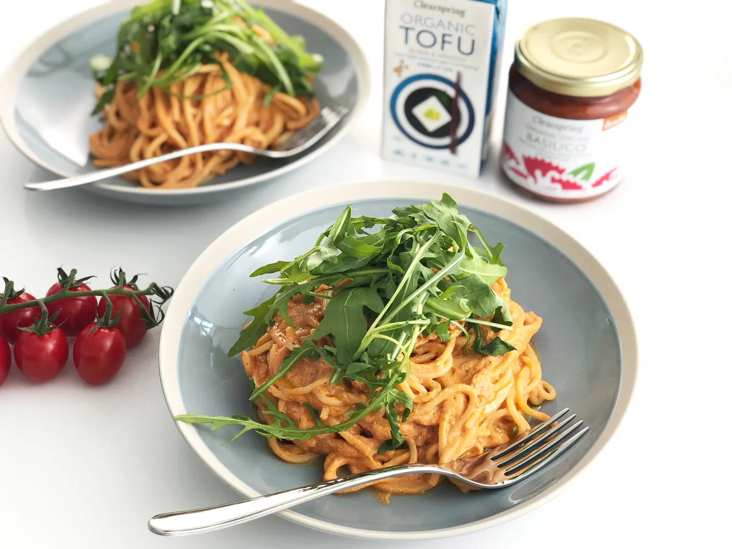 Clearspring Tofu Carbonara 300g Zwei Teller mit köstlicher Spaghetti Tofu Carbonara, garniert mit Rucola und zum Verzehr mit jeweils einer Gabel. Tomaten und ein Vorratsglas Basilikum Pesto und die 300g Vorratspackung Seiden Tofu