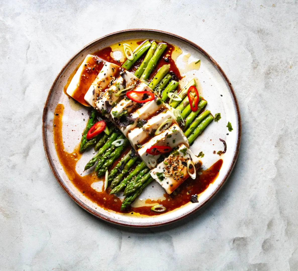 Clearspring Tofu Rezept 300g Auf einem Teller zubereiteter Tofu auf grünem Spargel, garniert mit Frühlingszwiebeln, Chilischoten und Sesam, mit brauner Sauce