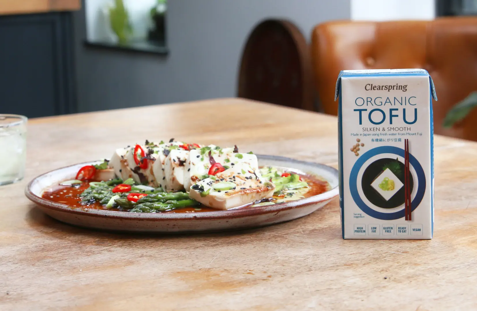 Clearspring Tofu Asparagus Salad 300g Auf einem Teller zubereiteter Tofu auf grünem Spargel, garniert mit Frühlingszwiebeln, Chilischoten und Sesam, mit Sauce. Nebenstehend Seiden Tofu in der 300g Vorratspackung