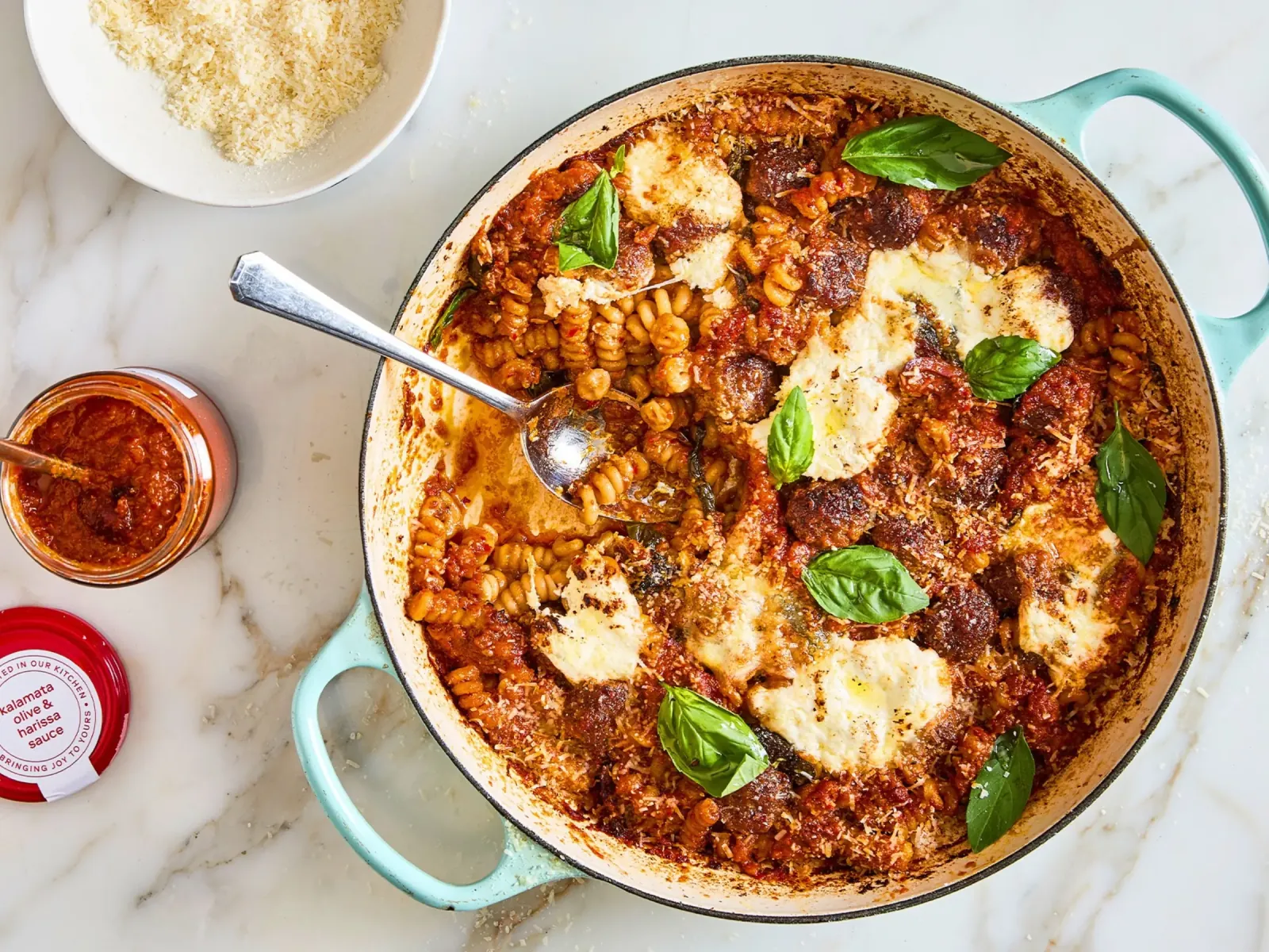 Pasta-Auflauf mit Fleischbällchen, Mozzarella und Basilikum im Schmortopf, daneben Ottolenghi Kalamata Olive & Harissa Sauce und geriebener Parmesan Pasta-Auflauf mit Fleischbällchen, Mozzarella und Basilikum im Schmortopf, daneben Ottolenghi Kalamata Olive & Harissa Sauce und geriebener Parmesan